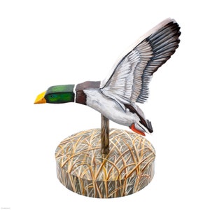 Op de afbeelding: Een houten eendenlokvogel met beschilderde details, geperst op een basis met een beschilderde afbeelding van riet. De eend is in vlucht met zijn vleugels uitgespreid.