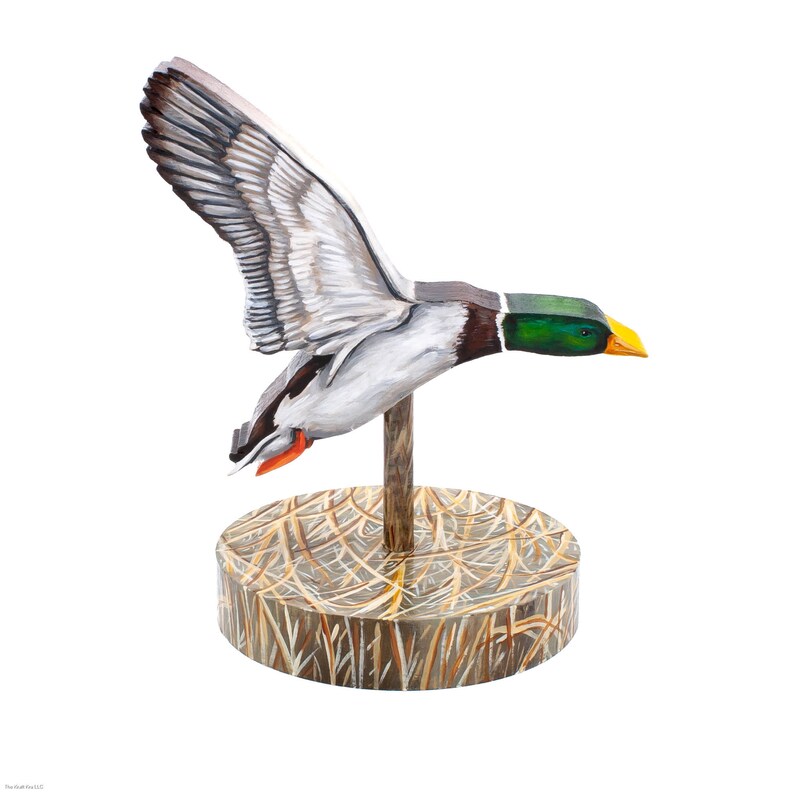 Op de afbeelding: Een houten eendenlokvogel met beschilderde details, geplaatst op een houten basis met een stroomachtige textuur. De eend is in vlucht met zijn vleugels uitgespreid en zijn kop gedraaid naar de zijkant.