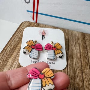 May include: A pair of novelty earrings and a charm shaped like a pencil and notebook bow. The earrings and charm feature a pink bow, a yellow pencil, and a notebook design. The earrings have gold-colored posts. The text "Slayed with Sarah" is visible.
