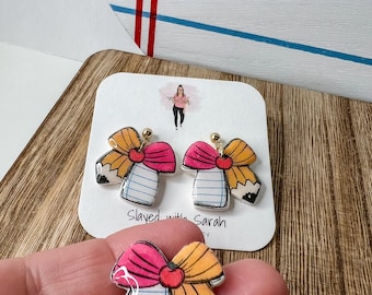 May include: A pair of novelty earrings and a charm shaped like a pencil and notebook bow. The earrings and charm feature a pink bow, a yellow pencil, and a notebook design. The earrings have gold-colored posts. The text "Slayed with Sarah" is visible.