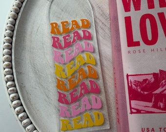 May include: Clear acrylic bookmark with the word "READ" in orange, pink, and yellow retro font. The bookmark is next to a pink book with the title "Wild Love". The items are on a white beaded tray.