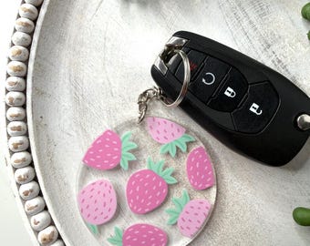May include: A clear acrylic keychain featuring a pattern of pink strawberries with green leaves. The keychain is attached to a silver keyring and a black car key fob. The design is on a white wooden tray.