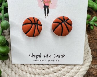 May include: A pair of handmade basketball stud earrings. The earrings are orange with black lines, resembling a basketball. The earrings are displayed on a white card with the text "Slayed with Sarah Handmade Jewelry".