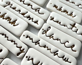 May include: White rectangular ceramic tags with various words in a cursive font, including "mama", "grandma", and "nurse". The words are in a contrasting brown color, and each tag has a small hole at the top.