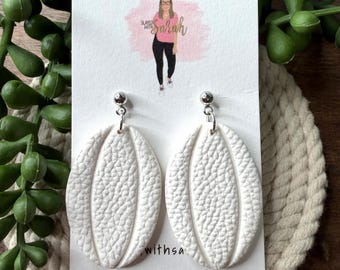 May include: A pair of white oval-shaped earrings with a textured, pebbled surface and a vertical center groove. The earrings are attached to a white display card featuring the text Slayed with Sarah HANDMADE JEWELRY and a graphic illustration.