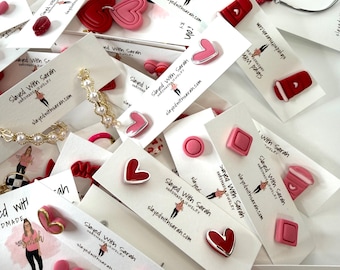 May include: A collection of handmade jewelry, including heart-shaped earrings, studs, and bracelets. The earrings come in various colors, including red, pink, and green. The jewelry is displayed on white cards with the text "Slayed With Sarah".