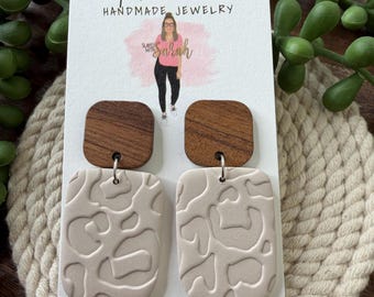 May include: Handmade earrings with a wood grain square top and a rectangular, beige, textured bottom. The earrings are displayed on a white card with the text "Slayed with Sarah Handmade Jewelry". Green plant sprigs are in the background.