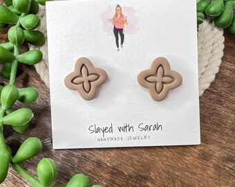 May include: A pair of brown clay earrings with a four-pointed star design. The earrings are displayed on a white card with the text "Slayed with Sarah HANDMADE JEWELRY".