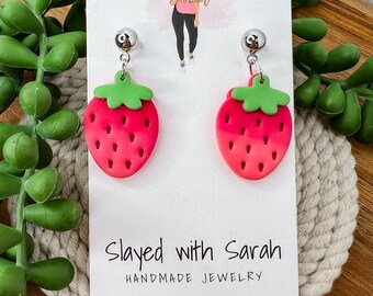 May include: A pair of red and green strawberry earrings with silver posts. The earrings are handmade and are displayed on a white card with the text "Slayed with Sarah Handmade Jewelry".
