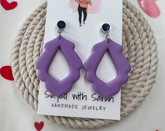 May include: Purple statement earrings with a unique, open design. The earrings are made of polymer clay and have a silver hook closure. The earrings are handmade and are a great addition to any jewelry collection.