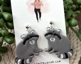 May include: A pair of gray raccoon earrings with black and white striped tails. The earrings are handmade and attached to a white card with the text "Slayed with Sarah HANDMADE JEWELRY" and the website address.