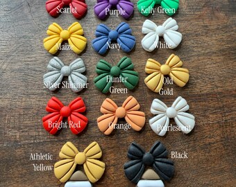 May include: A collection of bow-shaped stud earrings in various colors including yellow, royal blue, crimson, scarlet, purple, kelly green, maize, navy, white, silver shimmer, hunter green, old gold, bright red, orange, pearlescent, athletic yellow, and black.