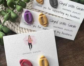 May include: Three cards with the text "Slayed With Sarah" and the words "Handmade Jewelry" on each card. Each card has two small football-shaped earrings on it. The earrings are in different colors: black, purple, gold, red, and yellow.