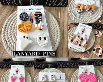 May include: A collection of Halloween-themed jewelry including lanyard pins featuring a spiderweb, pumpkin, ghost, and mummy, plus various earrings shaped like spiderwebs with bows, colorful hoops, pink skulls, ghosts with bows, and bright pink skeleton hands.