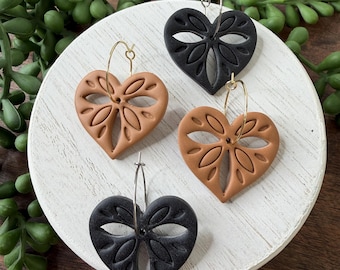 May include: Four heart-shaped earrings with a leaf-like design. Two are black, and two are a light brown color. The earrings are on a white, round surface with green plant accents. The earrings are on silver and gold hoops.