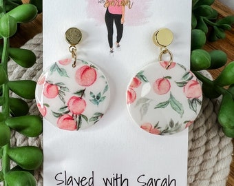May include: Peach-themed earrings with gold accents. The earrings are round and feature a white background with a peach pattern. The earrings are handmade and are labeled "Slayed with Sarah Handmade Jewelry".