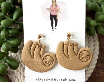 May include: Tan-colored sloth earrings with gold-colored ball studs. The earrings are displayed on a white card with the text "Slayed with Sarah Handmade Jewelry" and a website address. The background includes green foliage and a wooden surface.