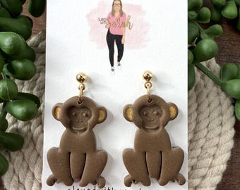 May include: Brown monkey-shaped earrings with gold accents. The earrings are displayed on a white card with the text "Slayed with Sarah Handmade Jewelry". The earrings are hanging from gold-colored posts.