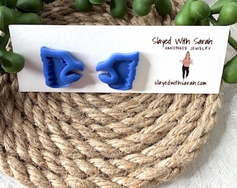 May include: A pair of blue handmade jewelry earrings in a unique design. The earrings are displayed on a white card with the text "Slayed With Sarah Handmade Jewelry" and the website "slayedwithsarah.com". The background includes a woven rope mat and green plant.