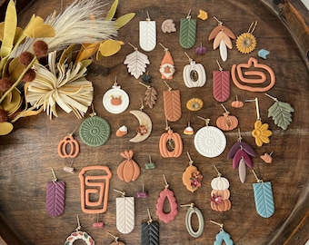 May include: A collection of fall-themed polymer clay earrings displayed on a wooden tray. The earrings feature various designs, including pumpkins, leaves, acorns, coffee cups, and wreaths. The earrings are in shades of orange, brown, green, and white.