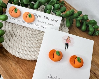 May include: A pair of peach-shaped earrings with green leaves. The earrings are on a white card with the text "Slayed With Sarah Handmade Jewelry".
