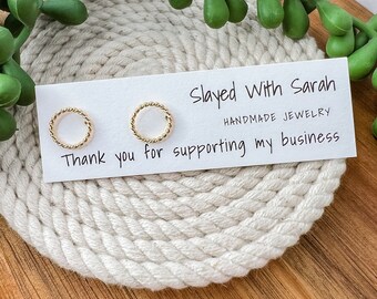 May include: A pair of gold-colored, twisted-rope design earrings. The earrings are displayed on a white card with the text "Slayed With Sarah Handmade Jewelry" and "Thank you for supporting my business". The card rests on a white rope coaster.