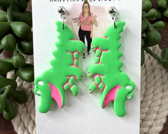 May include: A pair of handmade green dinosaur earrings with pink accents. The earrings are on a white card with the text "Slayed with Sarah HANDMADE JEWELRY" and the website "slayedwithsarah.com".