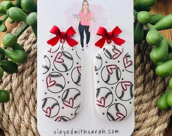 May include: White baseball-themed earrings with red heart accents and red bows. The earrings are on a white card with the text "Slayed with Sarah HANDMADE JEWELRY" and "slayedwithsarah.com". The background includes green plants and a woven rope.