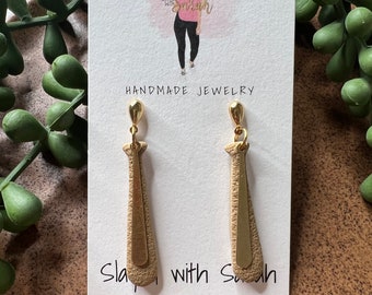 May include: A pair of gold-colored earrings with a textured, elongated design. The earrings are displayed on a white card with the text "Slayed with Sarah" and "Handmade Jewelry". The background includes greenery and a wooden surface.