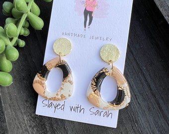 May include: A pair of gold and black geometric earrings with a white background. The earrings are shaped like a triangle with a rounded oval cutout. The earrings are handmade and have the text "Slayed with Sarah" on the card.