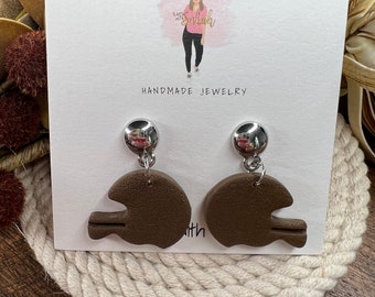 May include: A pair of brown clay earrings shaped like football helmets with silver metal hooks. The earrings are displayed on a white card with the text "Handmade Jewelry".