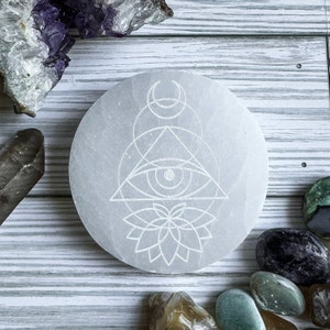 May include: White selenite charging plate with an etched design of an eye within a triangle, a flower, and a crescent moon.