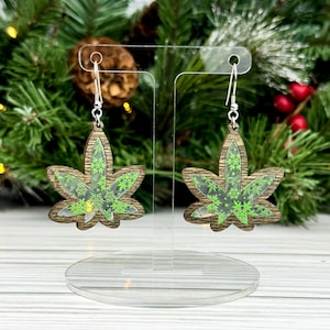 May include: A pair of wooden earrings shaped like marijuana leaves with a green snowflake pattern inside. The earrings are on a clear display stand.