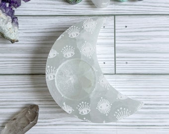 May include: White alabaster crescent moon candle holder with a carved eye design. The holder has a small indentation in the center for a tea light candle.