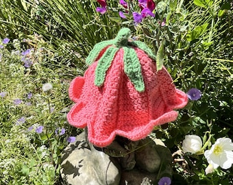 May include: A handmade crochet hat shaped like a pink flower with green leaves. The hat is set in a garden with purple and white flowers and green foliage. The hat's design is whimsical and colorful.