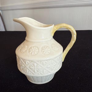 Puede incluir: Una jarra de crema de cerámica blanca con un asa amarilla. La jarra tiene un diseño de tejido de cesta y un patrón floral.