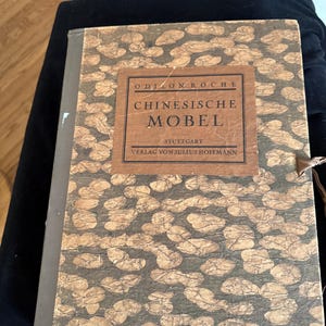 May include: A vintage book titled "Chinesische Möbel" with a brown and gold patterned cover. The book has a rectangular label with text in German. The book is closed and has a string tie.