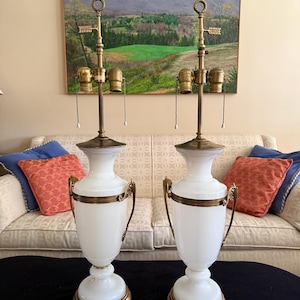 Può includere: Coppia di lampade da tavolo in vetro bianco con dettagli in ottone. Ogni lampada presenta una base bombata, un corpo cilindrico e una parte superiore in ottone con catenella. Le lampade hanno manici decorativi in ottone.