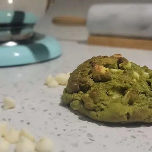 May include: A green matcha cookie with white chocolate chips. The cookie is on a white countertop with white chocolate chips scattered around it.