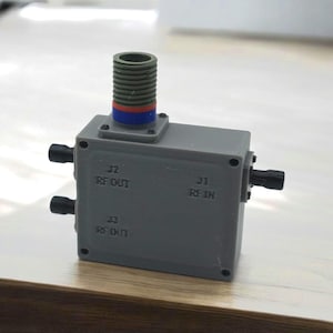 以下が含まれることがあります： J1、J2、J3 とラベル付けされた 3 つのポートを持つ灰色のボックス。J1 は「RF IN」、J2 は「RF OUT」、J3 は「RF OUT」とラベル付けされています。赤、青、白のバンドが付いた灰色の円筒形コネクタがボックスの上部に接続されています。