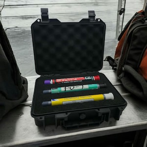 May include: A black, hard-shell case containing three medical injectors. The injectors have coloured barrels: red, green, and yellow, with printed text. The case is open, revealing a foam interior.