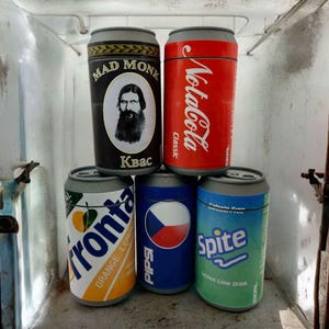 May include: Five aluminium cans with different designs. The cans are arranged in a row, with the first can on the left showing a man with a beard and the text "Mad Monk Kbac". The second can shows the text "Coca-Cola Classic". The third can shows the text "Fronta Orange Lemon". The fourth can shows the text "Pips" and a Czech flag. The fifth can shows the text "Spite Lemon Lime Drink".