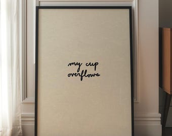 May include: A framed print with the words "my cup overflows" in black script. The print has a cream-colored background and a black frame. The print is leaning against a white wall next to a wooden cabinet and a white lamp.
