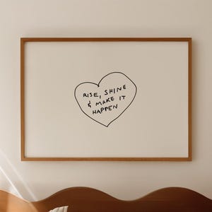 Op de afbeelding: Een ingelijste print met de woorden "Rise, Shine & Make It Happen" in een hartvorm. De print zit in een houten lijst en hangt boven een bed met gestreepte beddengoed en een houten hoofdbord.