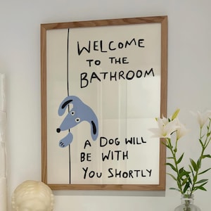 May include: A framed print with the text "WELCOME TO THE BATHROOM" and a cartoon dog illustration. The print is in a bathroom setting, with a stack of toilet rolls, a bust, a candle, and a vase of flowers.