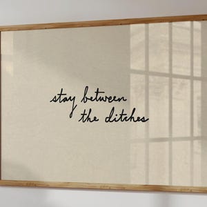 Puede incluir: Impresión enmarcada con la frase "stay between the ditches" en escritura negra sobre fondo beige. El marco de madera resalta el texto. Una decoración de pared sencilla y elegante para el hogar.