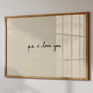 Puede incluir: Una impresión enmarcada con el texto manuscrito "p.s. i love you" en escritura negra. La impresión tiene un fondo beige y un marco de madera natural. La imagen está montada en una pared blanca.
