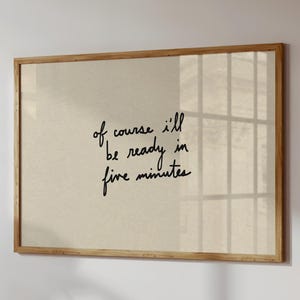 May include: A framed print with the handwritten text "of course I'll be ready in five minutes" in black script. The print has a beige background and a light brown wooden frame. The artwork is mounted on a white wall.
