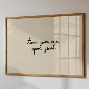 May include: A framed print with the handwritten text "turn your eyes upon jesus" in black script. The print has a beige background and a light brown wooden frame. The artwork is hanging on a white wall.
