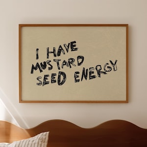 May include: A framed print with the text "I HAVE MUSTARD SEED ENERGY" in black lettering. The print is displayed above a bed with a wooden headboard and striped bedding. The overall color scheme is neutral with warm tones.